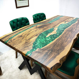 A dining table with a Black Walnut Wood top and pearl green epoxy river design, surrounded by green upholstered chairs in a room with a white wall and wood flooring.
