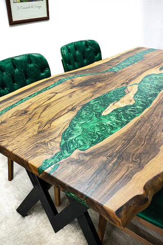 A dining table with a Black Walnut Wood top and pearl green epoxy river design, surrounded by green upholstered chairs in a room with a white wall and wood flooring.